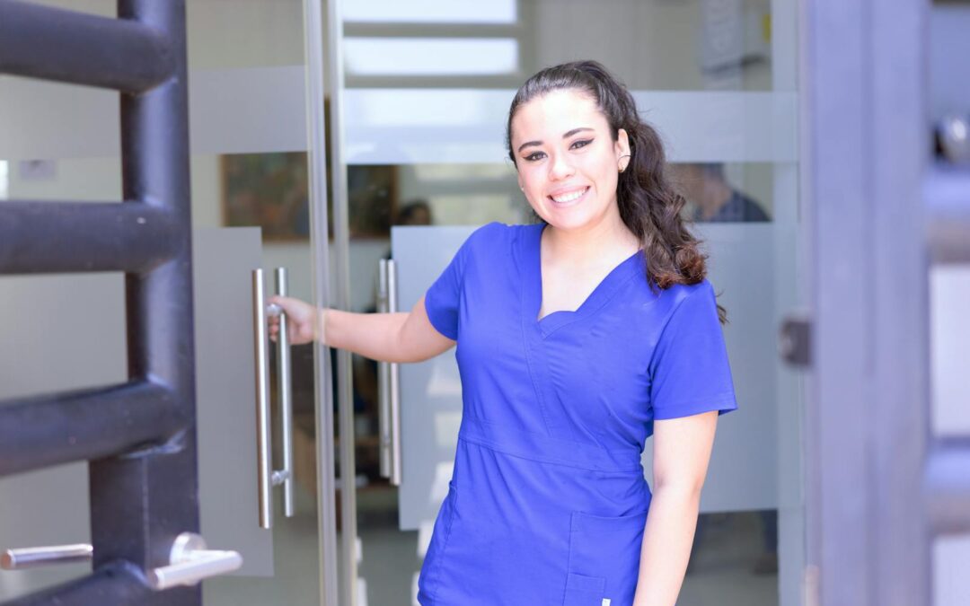 Smiling female nurse in blue scrubs opens a glass door in a healthcare setting.